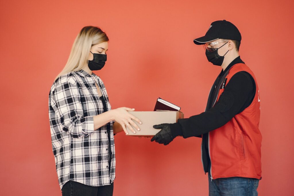 Side view of adult delivery man in protective mask and goggles with gloves giving carton parcel to woman in checkered shirt and medical mask isolated on red background illustrating concept of safety delivery during quarantine
